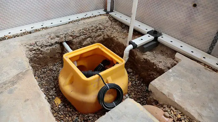 A yellow sump basin installed in the ground, surrounded by gravel and connected to drainage pipes