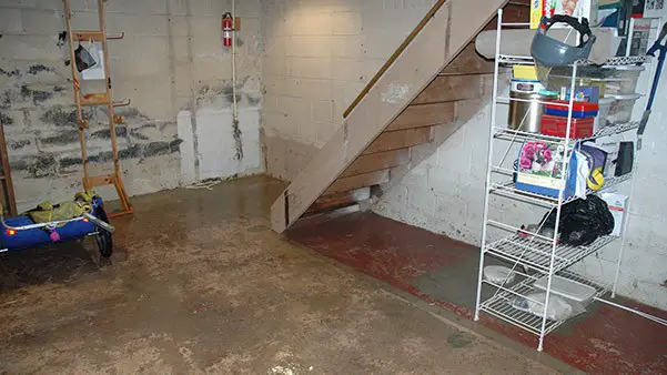 A basement area with water on the floor, indicating a flood or leakage issue. A wooden staircase with a simple railing leads upwards out of the basement
